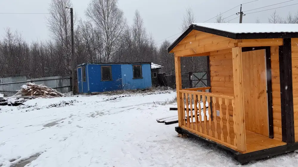 Готовая мобильная баня-бытовка 2,3 на 7 метров с крыльцом и балясинами на участке с другой бытовкой из профлиста на заднем плане