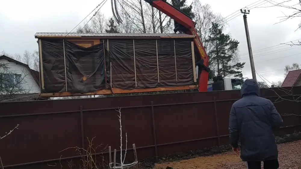 Разгрузка мобильной бани автокраном через забор участка: баня в плёнке поднимается стрелой крана в сложных условиях заезда