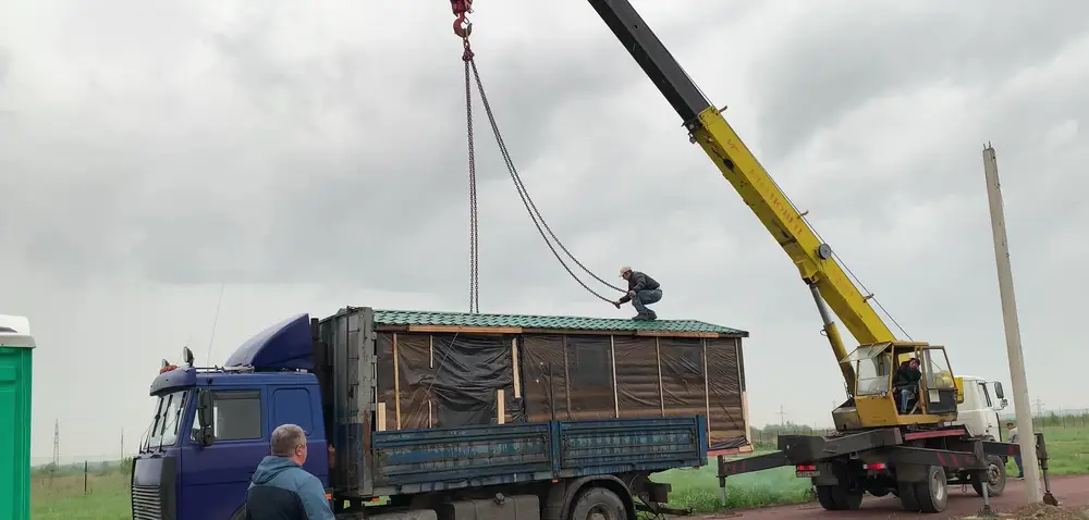 Водитель на крыше бани под стропами автокрана закрепляет цепной паук, работа на металлочерепице во время дождя